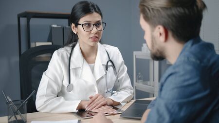 Female doctor listens to male patient complaints asks questions sitting at desk working in hospitalの写真素材