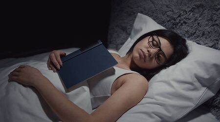 Portrait of young lady with glasses lying on back in bed fallen asleep while reading book, side viewの写真素材