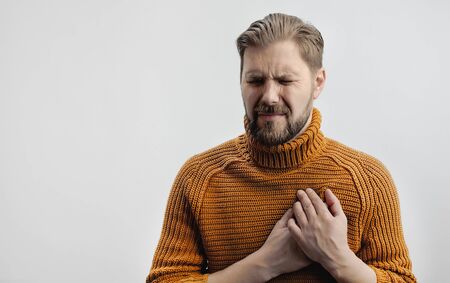 Portrait of grimacing bearded man placing hands on chest suffering from heartache, isolated