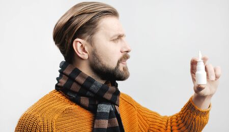 Adult man in scarf and yellow sweater looking at nasal spray medicine, side view, white backgroundの写真素材