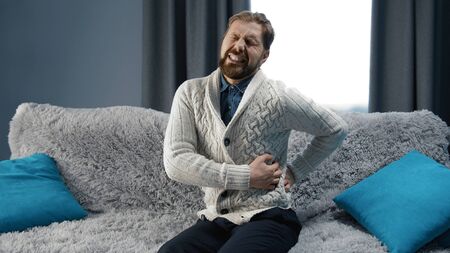Mature man with painful expression sitting on sofa touching waist suffering from back pain at home