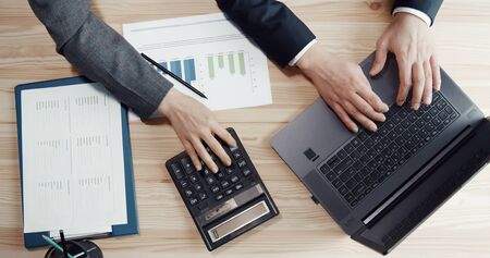 Cropped image of three hands during accounting process using laptop and calculator, top view