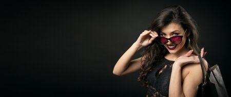 Glad brunette holding shopping bags on shoulder looking camera over sunglasses, black copyspace