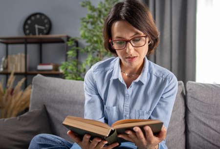 Chrming woman in eyeglasses and blue shirt getting interested while reading new book at home....