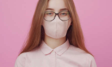 Cropped headshot of young lady in glasses wearing facemask looking at camera, isolated pink backgroundの写真素材