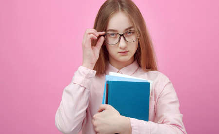 studio shot of young lady holding books touching glasses posing to camera isolated on pink backgroundの写真素材