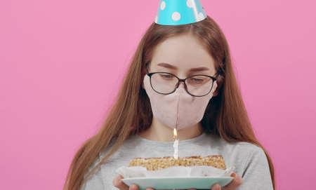 Headshot of girl with facemask holding birthday cake looking at burning candle, isolated on pinkの写真素材