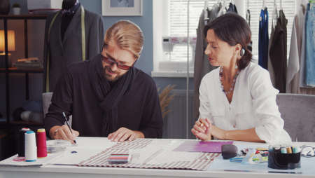 Two fashion designers discussing ideas sketching on paper sitting at desk in sewing atelierの写真素材