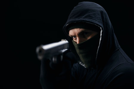 Angry gangster in hood, mask and gloves holding weapon in hands over black background. Bandit aiming with gun in studio.の写真素材