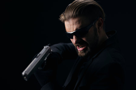 Aggressive bearded man in glasses, suit and gloves threatening someone with real gun. Isolated over black background. Concept of danger.の写真素材