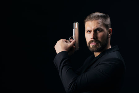 Aggressive bearded bandit in black suit standing over black background with real weapon in hands....
