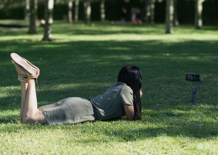 A woman is laying on the grass and taking a picture with a camera. The scene is peaceful and relaxing, with the woman enjoying the outdoors and capturing a moment in timeの写真素材