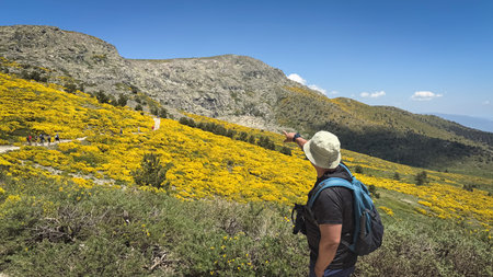 A man wearing a hat and a backpack is pointing at a mountain. The man is standing on a hill with a...