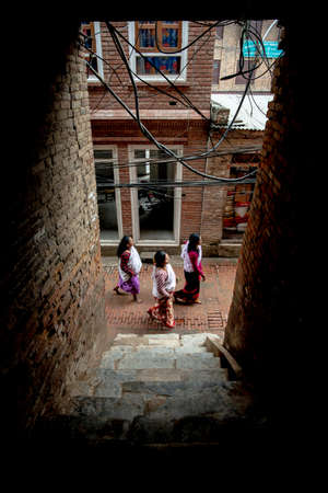 Bhaktapur, Nepal - April 5, 2019: Three women wearing traditional clothes walking in a narrow street, seen from a dark porch and taken in an overcast morning in the historical center of Bhaktapurのeditorial素材