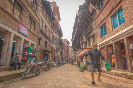 Bhaktapur, Nepal - April 5, 2019: Old man porting plastic cans walking down a narrow street taken during a partly sunny afternoon in the historical center of Bhaktapurのeditorial素材