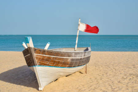 Disused traditional fishing boat and Qatar flag sitting on a sandy beach in front of the sea taken in the afternoon in Souk Wakrah, Qatarの写真素材