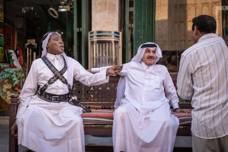 Doha, Qatar - 3 Dec 2016: Two men from Qatar are discussing on a bench in front f an Indian man. Taken in Souq Wakif, Dohaのeditorial素材