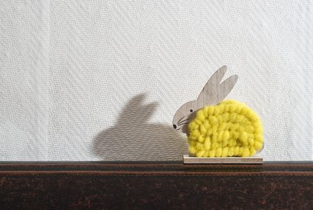 Yellow easter bunny in front of white wall in a room. Bunny decoration and white wallpaper background. Shadow of rabbit on the wall. Wooden bunny figure shape and yellow yarn. Minimalist concept.の写真素材