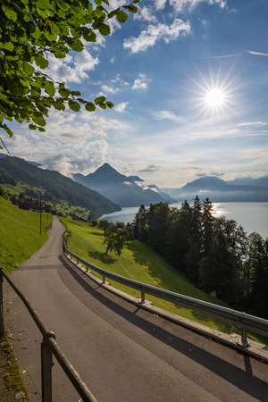 Picturesque road running away from the camera along an alpine lake with a bright sunburst and distant mountain peaks in summerの写真素材