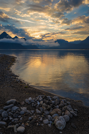 Tranquil colorful sunrise over a calm lake or sea with pebbles on the shoreline in the foreground and the rising sun over the mountains reflected on the surface of the still waterの写真素材