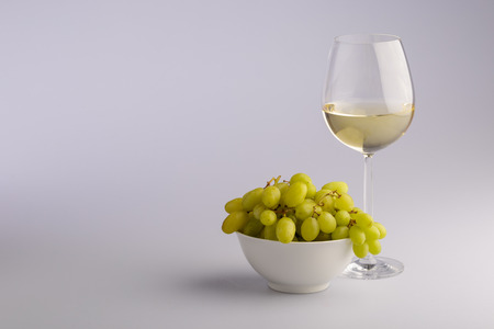 Elegant glass of white wine with a bunch of fresh green grapes in a bowl in a concept of wine making and viticulture on a grey background with copy spaceの写真素材