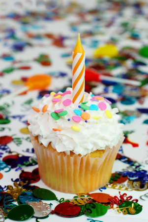 Celebration cupcake with candle and sprinkles.  Confetti and suckers in background.