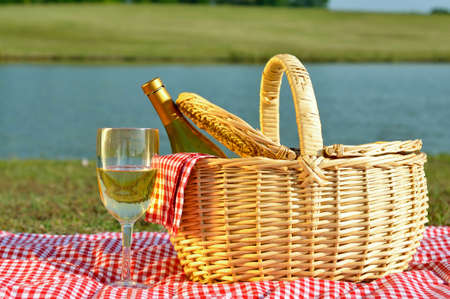Bottle of white wine in picnic basket with glass of wine beside it.  Red gingham blanket and napkin with lake in background.の写真素材