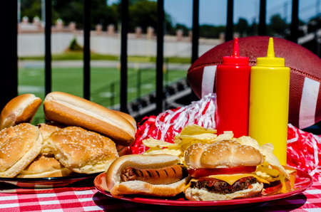 Football party with cheeseburger, hot dog, potato chips, pom poms, buns, and football   Football field in background の写真素材