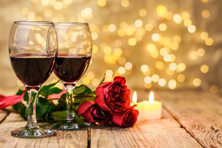 Valentine's Day concept. Two glasses of wine, red roses and burning candles on a rustic wooden table in front of a blurred luminous background with copy space for text. Selective focus.の写真素材