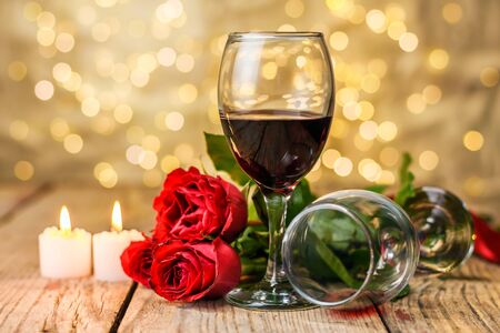 Valentine's Day still life. Two glasses of wine, red roses and burning candles on a rustic wooden table in front of a blurred luminous background. Selective focus.の写真素材