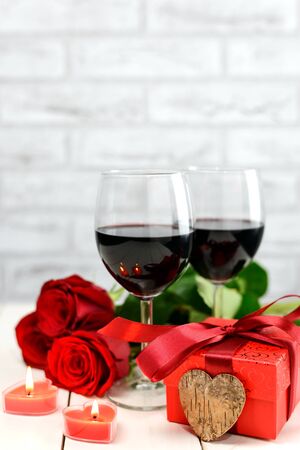 Valentine's Day concept. Two glasses of wine, red roses, gift box, wooden heart and burning candles on a white wooden table. Selective focus.の写真素材