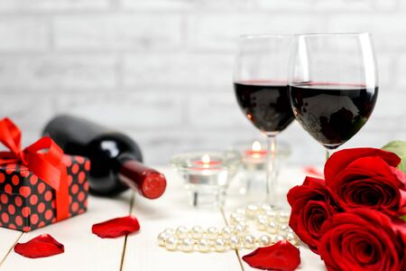 Valentine's Day concept. Two glasses of wine, red roses, gift, string of pearls, rose petals, wine bottle and burning candles on a white wooden table. Selective focus.の写真素材