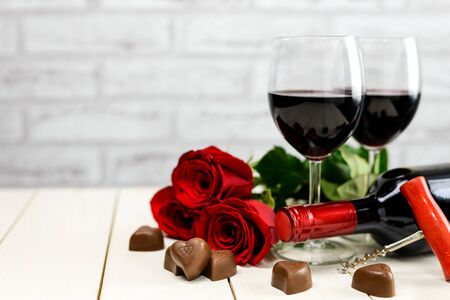Valentine's Day concept. Two glasses of wine, wine bottle, corkscrew, red roses and chocolate hearts on a white wooden table with copy space for text. Selective focus.の写真素材