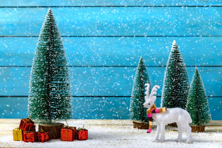 Miniature reindeer standing near fir trees and Christmas gifts under falling snow on wooden table. Concept of celebrating Christmas and New Year.の写真素材