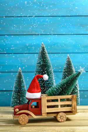 Christmas holiday celebration concept. Christmas tree on a toy truck with a Santa hat next to fir trees while it is snowing on a wooden table.の写真素材