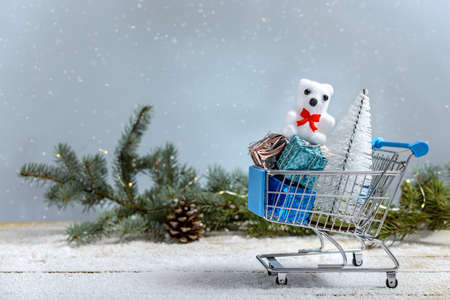 Christmas shopping concept. Christmas tree, Teddy bear and gifts in a shopping cart next to branch fir tree while it is snowing on a wooden table.の写真素材