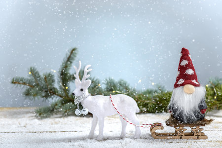 Sleigh with Christmas gnome and white reindeer next to fir branch while it is snowing on a wooden table. Christmas and New Year concept.の写真素材