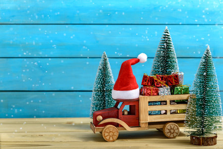 Christmas gifts on a to truck with a Santa hat next to fir trees while it is snowing on a wooden table.の写真素材