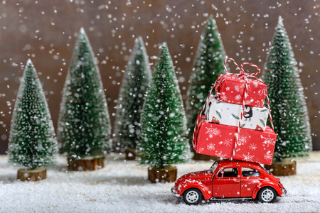 Red toy car with a Christmas gifts on the roof next to fir trees while it is snowing on a wooden table. Concept of celebrating Christmas and New Year.の写真素材