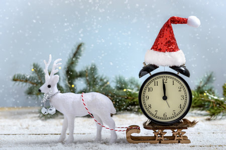 White reindeer pulls clock with Santa hat on a sled next to fir branch while it is snowing on a wooden table. Christmas and New Year concept.の写真素材