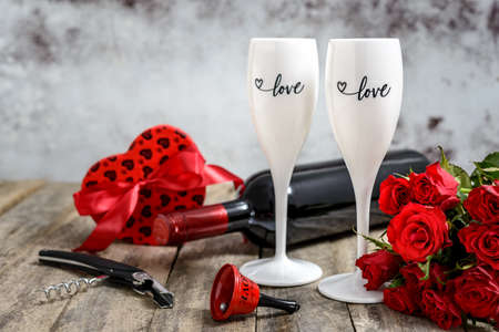 Valentine's Day concept. Two glasses of wine, red roses, gift, bell, wine bottle and corkscrew on a rustic wooden table. Selective focus.の写真素材