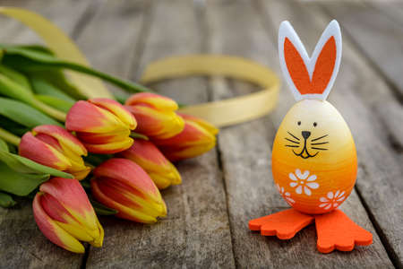 Easter egg with cute bunny face and rabbit ears next to orange-yellow tulips on a rustic wooden table. Easter concept.の写真素材