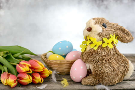 Funny bunny made of straw with Easter eggs painted, feathers and orange-yellow tulips on wooden background. Easter concept.の写真素材