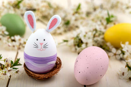 Easter egg with cute bunny face and rabbit ears next to blossomed spring flowers and colorful eggs on a wooden table. Easter concept.の写真素材