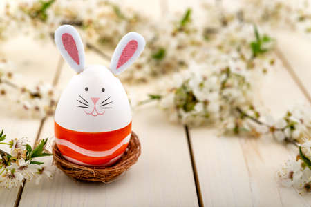 Easter egg with cute bunny face and rabbit ears next to blossomed spring flowers on a wooden table....
