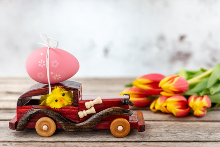 A wooden toy car with an Easter egg on the roof and a chicken driver next to a tulip bouquet on a...