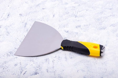 Steel trowel scraper or spatula with black-yellow rubberized handle on white-grey background.