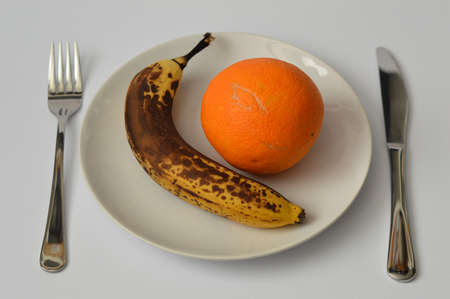 Overripe banana and orange on the plate with fork and knife on light gray backgroundの写真素材