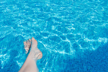 A serene scene with woman's feet submerged in a clear blue pool, capturing relaxation and summer...