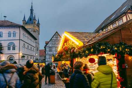 A lively Christmas market scene in a medieval town in Bad-Wimpfen, Germany. Warmly lit winter holiday stalls, garlands, and cheerful visitors. Half-timbered houses and a tower as historic backdrop.の写真素材